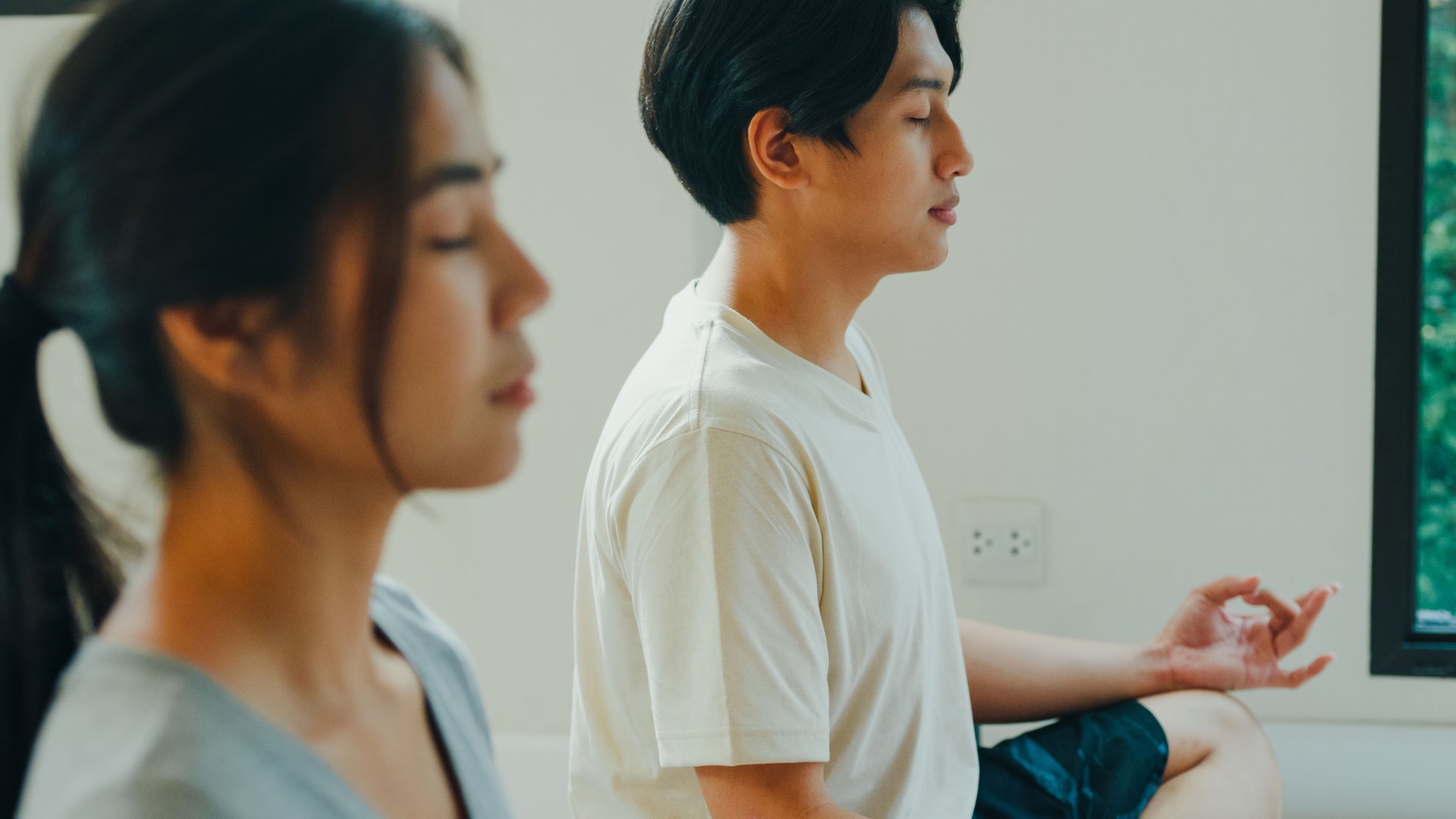 Closeup of cheerful young Asian serene couple meditates in the lotus position in a peaceful home setting, with a laptop nearby for guided meditation. Enjoying home fitness workout.